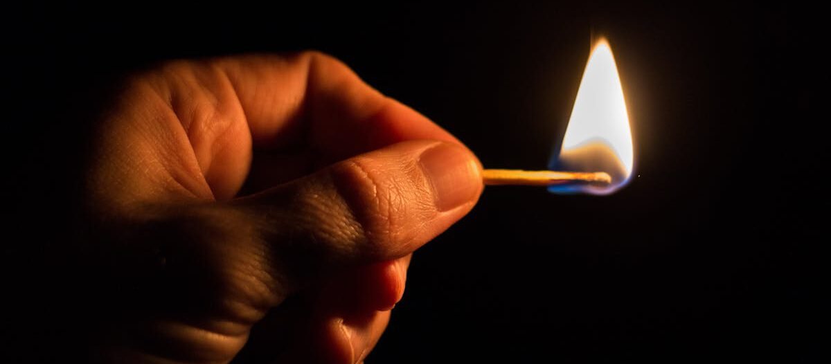 Close-up of a hand holding a lit match against a dark background, highlighting the flame.
