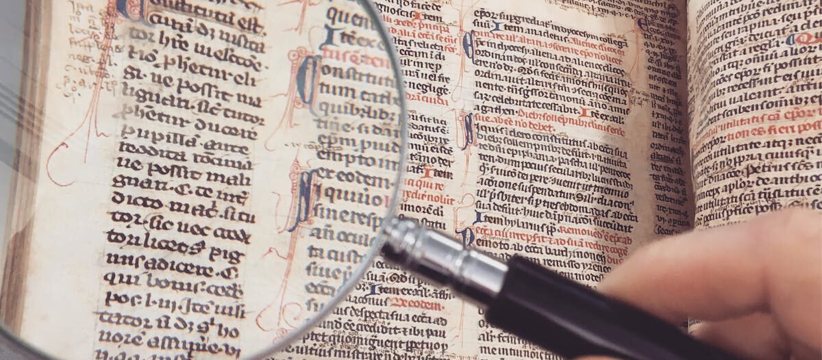 A hand holding a magnifying glass over an antique book page, revealing intricate text and design.