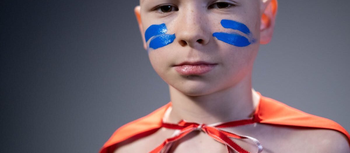 A young boy with blue face paint wearing a red superhero cape. Studio portrait.