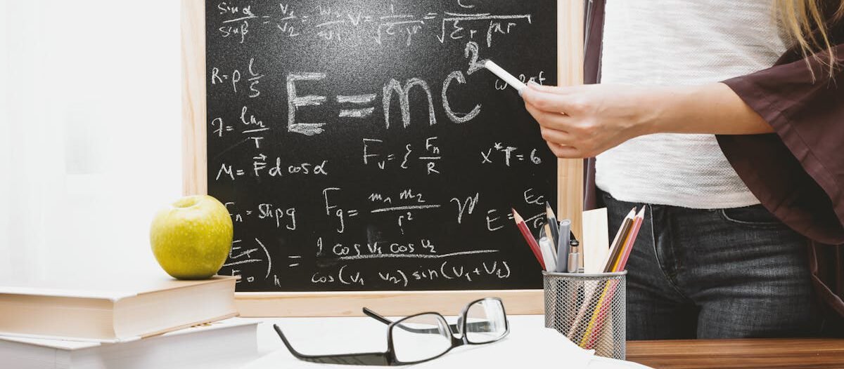 Woman writing physics equations on a blackboard with books and an apple on the desk.